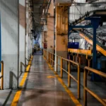 Interior pabrik modern dengan mesin produksi dan sistem konveyor yang beroperasi, menggambarkan perencanaan produksi manufaktur